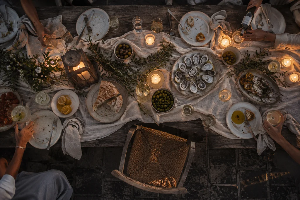 Overhead view of a candlelit coastal dinner table with oysters, olives, bread, wine, and shared plates after a relaxed evening meal.