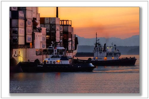 Crowley's Athena and Guard working a Matson container ship in Puget Sound at sunset — orange sky, mountain silhouettes, two t...