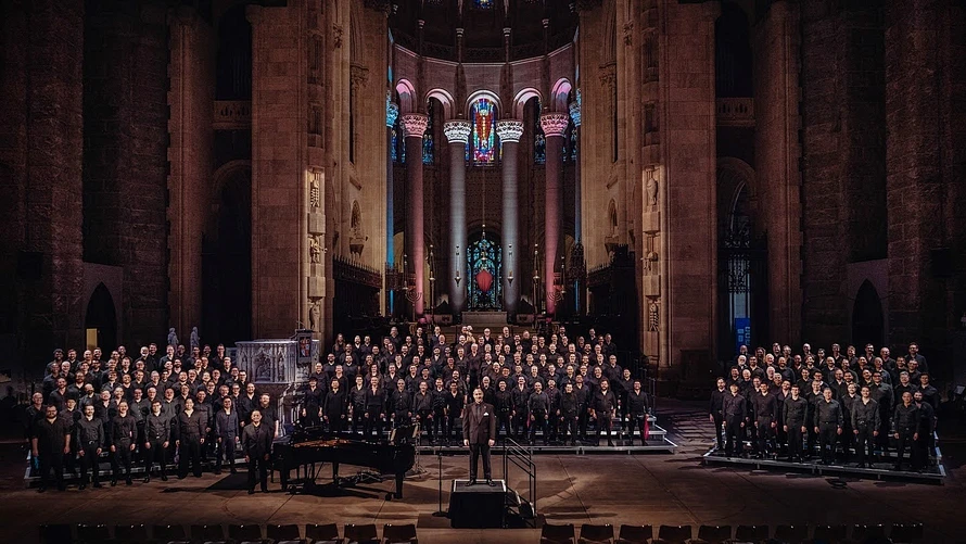 New York City Gay Men's Chorus standing on choral risers in the Cathedral of St. John the Divine. The background is up-lit with the light blue, pink, and white of the Trans flag. Photo credit: Mikey Gulcicek @mikeygulci  