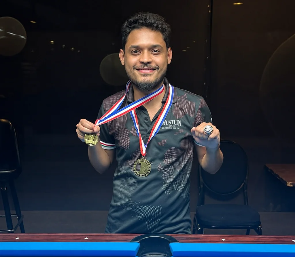 A smiling pool player holds up his medals beside a blue pool table.