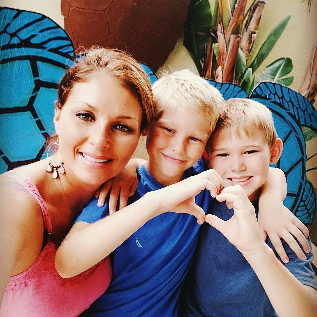 Woman smiling with two young boys in a family photo, with one child forming a heart shape with his hands against a colorful tropical mural background.