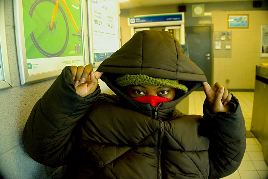 Artist Love, Builder of Worlds with their brown hoodie up in a train station wearing a red N95 mask looking directly into the camera.