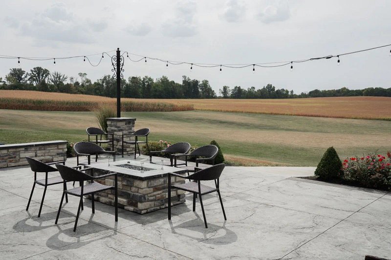 A stone patio with outdoor chairs, a fire table, and string lights overlooking open farmland and distant trees