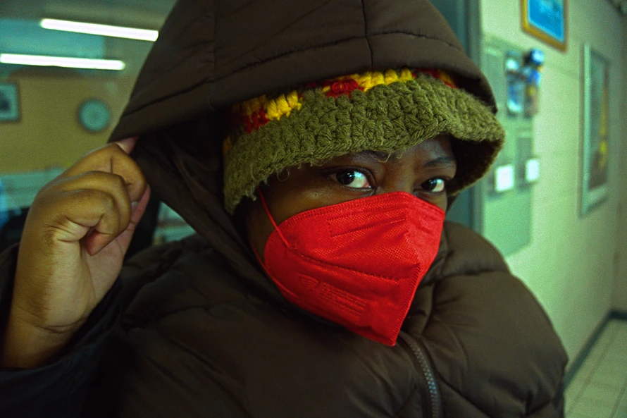 Image description : Love, Builder of Worlds with their brown hoodie up in a train station wearing a red N95 mask looking directly into the camera.