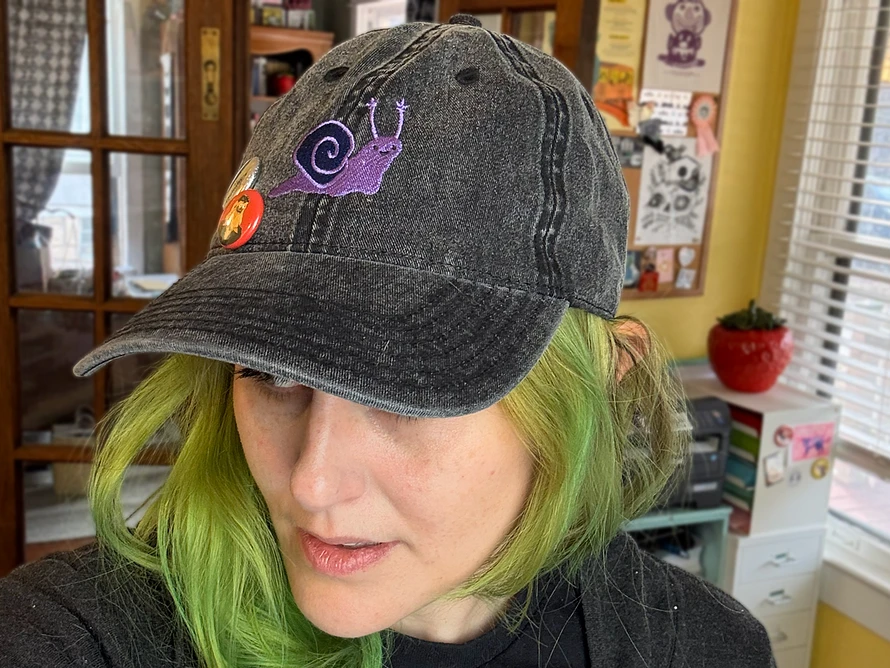 A white person with green hair wearing a dark grey hat embroidered with a purple snail.