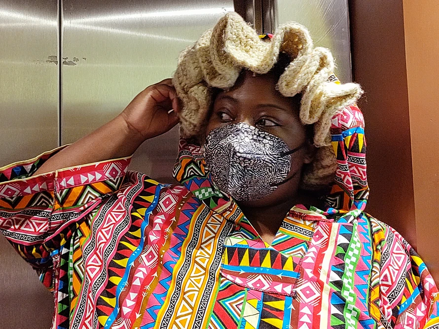 Love, Builder of Worlds, a West African person wearing bright colourful West Africa clothes, and a grey animal print FFP2 mask and a swirly beige knitted hat in front of elevator doors. They are looking away.