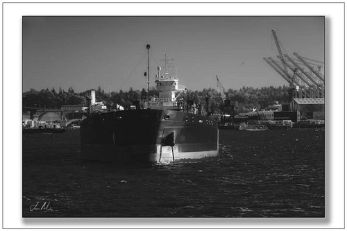 Crowley's ATB riding at anchor in Elliott Bay — bow-on and squared up, shipyard and container cranes framing the background. ...