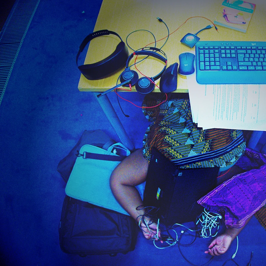 Single Artwork description: An African person on the floor under a desk appearing exhausted and overwhelmed. He is wearing traditional African material, surrounded by office items: a laptop covering his face, theres wires to the right of him, a laptop bag to the left of him and on the office table theres an air purifier necklace, headphones, an ergonomic mouse, a regular mouse, a wireless keyboard, papers, a book and a wireless HDMI transmitter. The image is overall is a bluey hue.
