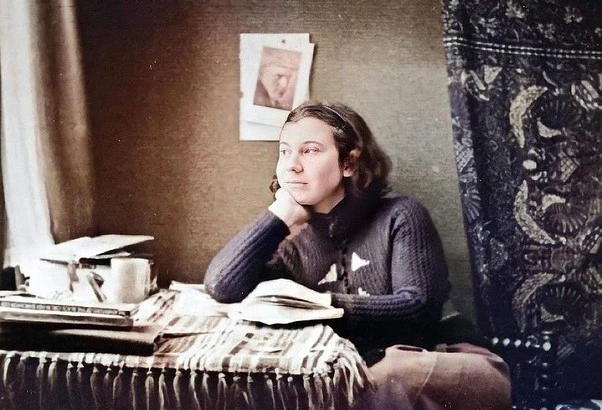 Etty Hillesum sits at a table looking pensively out of a window while her hand rests on an open books.